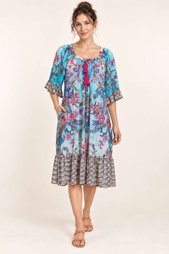 Turquoise Rhapsody Frill Dress, a blue floral and paisley viscose rayon dress, studio front view with a hand in the side pocket.