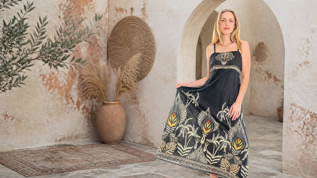Malie Mood Dress, a black strappy maxi dress with gold and cream palm-leaf print, worn in a sunlit courtyard